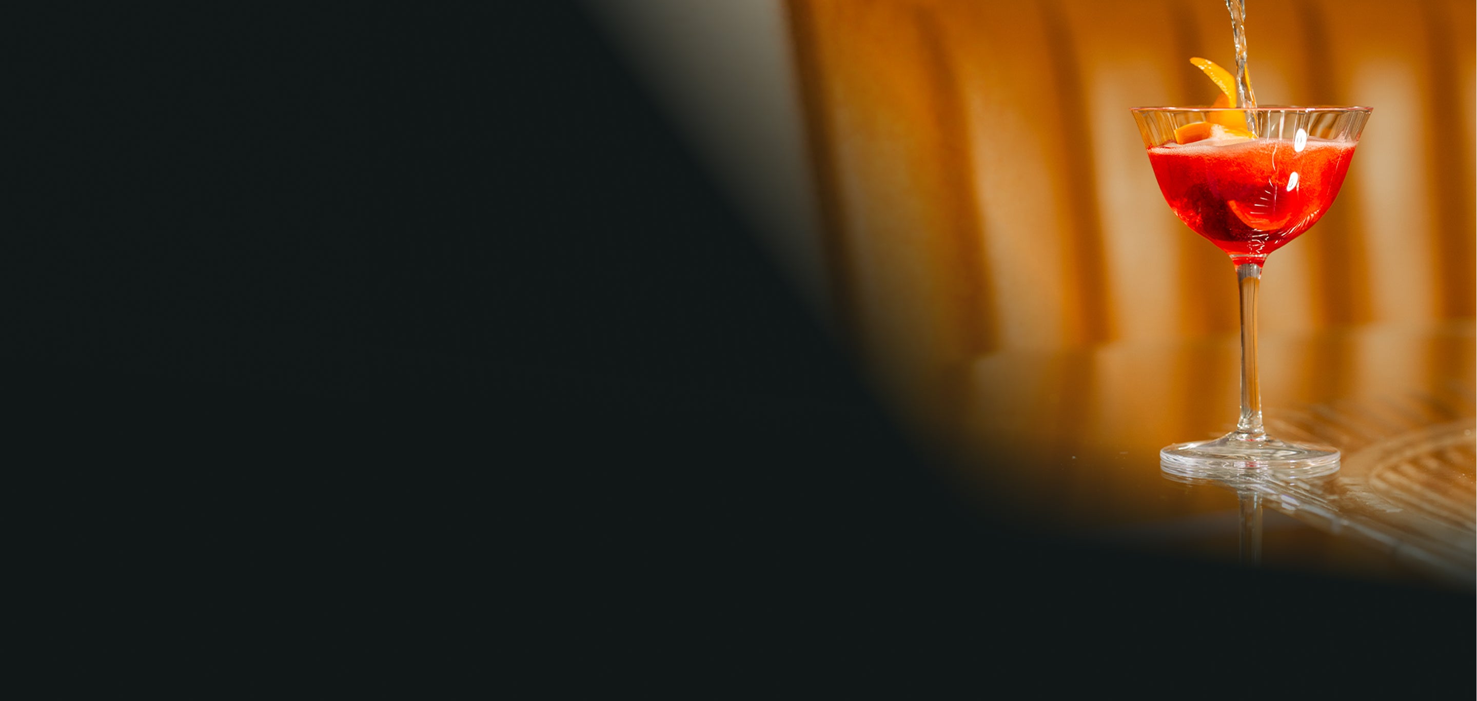A stemmed cocktail glass filled with a bright red drink and garnished with an orange peel, set against a warm golden-brown blurred background with a dark foreground shadow.